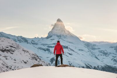 Voyageur en veste rouge debout sur une crête enneigée contemplant le mont Cervin (Matterhorn) illuminé par le soleil levant dans les Alpes.